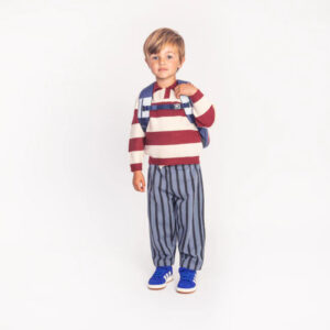 Young child standing against a white background, wearing a red-and-white striped sweater, blue striped pants, blue sneakers, and a blue backpack.