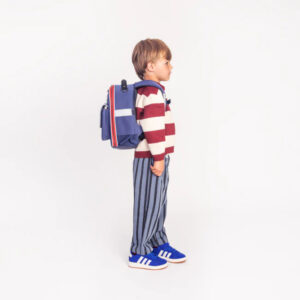 Side view of a young boy wearing a blue backpack, burgundy-and-white striped sweater, and blue striped pants, facing right.