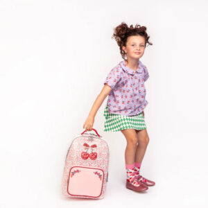 Young girl posing in a patterned purple top and green diamond skirt, holding a pink floral backpack with cherry design beside her.