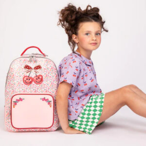 Young girl sits on the floor beside a pink floral backpack with red handles and cherries design.