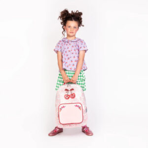 Young girl with two puffy buns, wearing a purple top and green checkered skirt, holding a pink floral backpack.