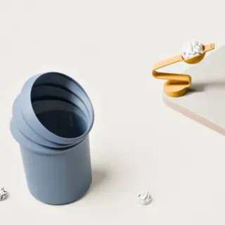 Blue modular cup tipped open on a pale surface, with crumpled paper balls and a yellow spiral paperweight on a wooden block.