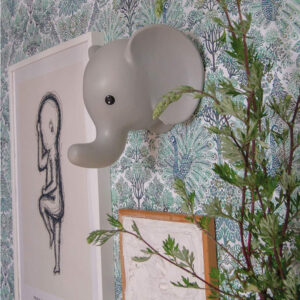 Grey elephant head wall lamp mounted above a framed line drawing, with leafy branches and botanical wallpaper in the background.