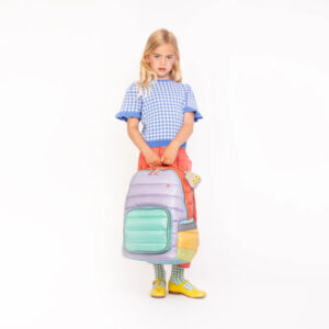 Girl standing with a pastel colorblock backpack, wearing a blue gingham top and red pants, looking at the camera.