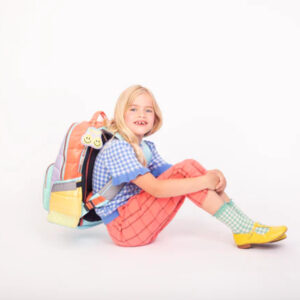 Smiling young girl with a multicolored backpack sitting on the floor, wearing a blue checkered top and pink pants.