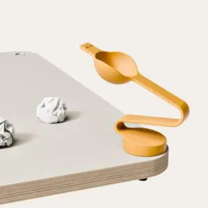 Orange adjustable clamp-like desk accessory attached to the edge of a light wood table, featuring a curved arm and scoop, with two crumpled papers on the surface nearby.