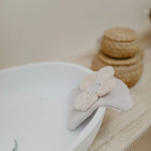 Beige fabric flower attached to the rim of a white ceramic bowl, with woven baskets in the background.