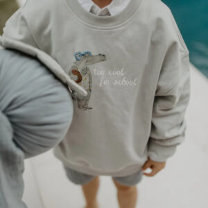 Child wearing a light gray sweatshirt with a cartoon crocodile and the text 'too cool for school'.