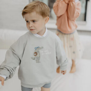 Young boy in a light gray sweatshirt with a cartoon dinosaur, walking indoors.
