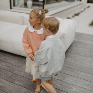 Two young children holding hands on a wooden deck, girl in peach sweater with white collar and skirt, boy in a gray jacket.