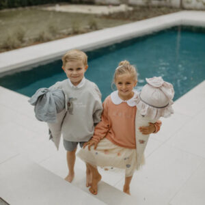 Two smiling children stand barefoot on poolside steps, each holding a large stuffed accessory and looking at the camera.