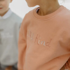 Close-up of a peach sweatshirt with pale embroidered text reading 'Be kind' across the chest.