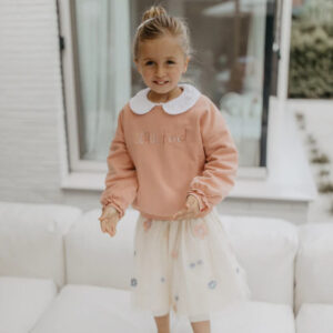 Young girl standing on a white sofa, wearing a peach sweater with a white collar and a light tulle skirt, smiling at the camera.