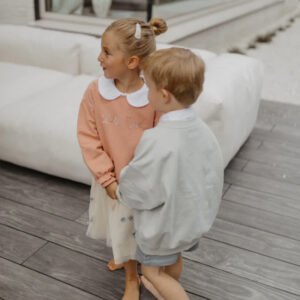 Two young children holding hands on a wooden deck; the girl wears a peach dress with a white collar, the boy wears a light gray jacket.