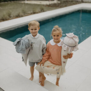 Two smiling children stand on pool deck barefoot, each carrying a bundled blanket and soft toy.
