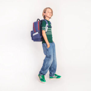 Young boy with a blue backpack, green striped shirt, loose jeans, and bright green sneakers standing against a white background.