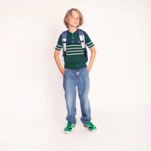 Child wearing a green striped polo, loose blue jeans, green sneakers, and a purple backpack, standing with hands in pockets.