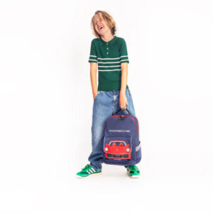 Child wearing a green polo and jeans smiles while holding a blue Porsche backpack with a red car graphic.