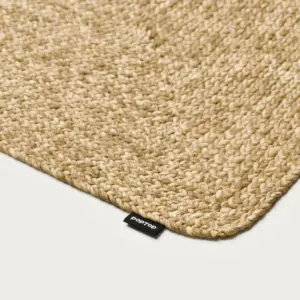 Close-up of a beige braided jute rug with a small black brand tag in the corner on a white floor.