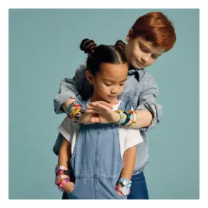 Two kids, a girl in denim overalls in front and a boy behind, embracing her wrists with colorful bracelets and watches; teal backdrop.