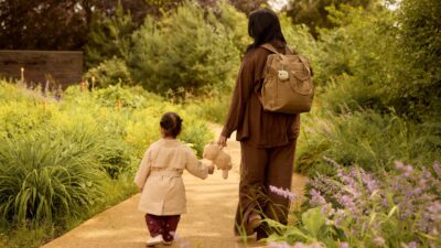 Mutter und Kleinkind mit Kuscheltier gehen über einen Gartenweg – Jollein Babyausstattung